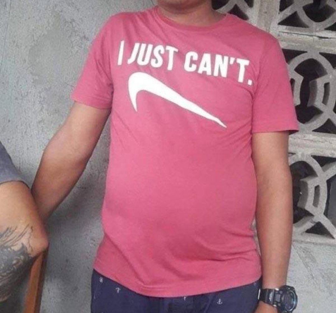 A person wearing a red T-shirt that parodies the Nike logo, reading "I JUST CAN'T." with a swoosh underneath, stands next to a concrete wall with patterned blocks.