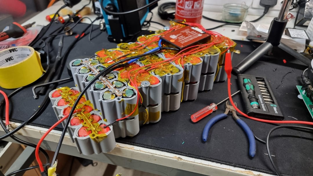 A homemade battery pack with multiple cylindrical cells connected by wires sits on a cluttered workbench surrounded by tools, electrical tape, and electronic components.