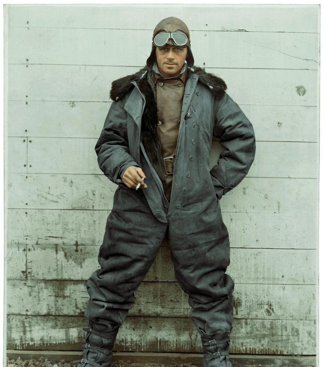 A man in a vintage aviator outfit, including goggles, fur-lined coat, and heavy boots, stands against a weathered wooden wall, holding a cigarette in his right hand.