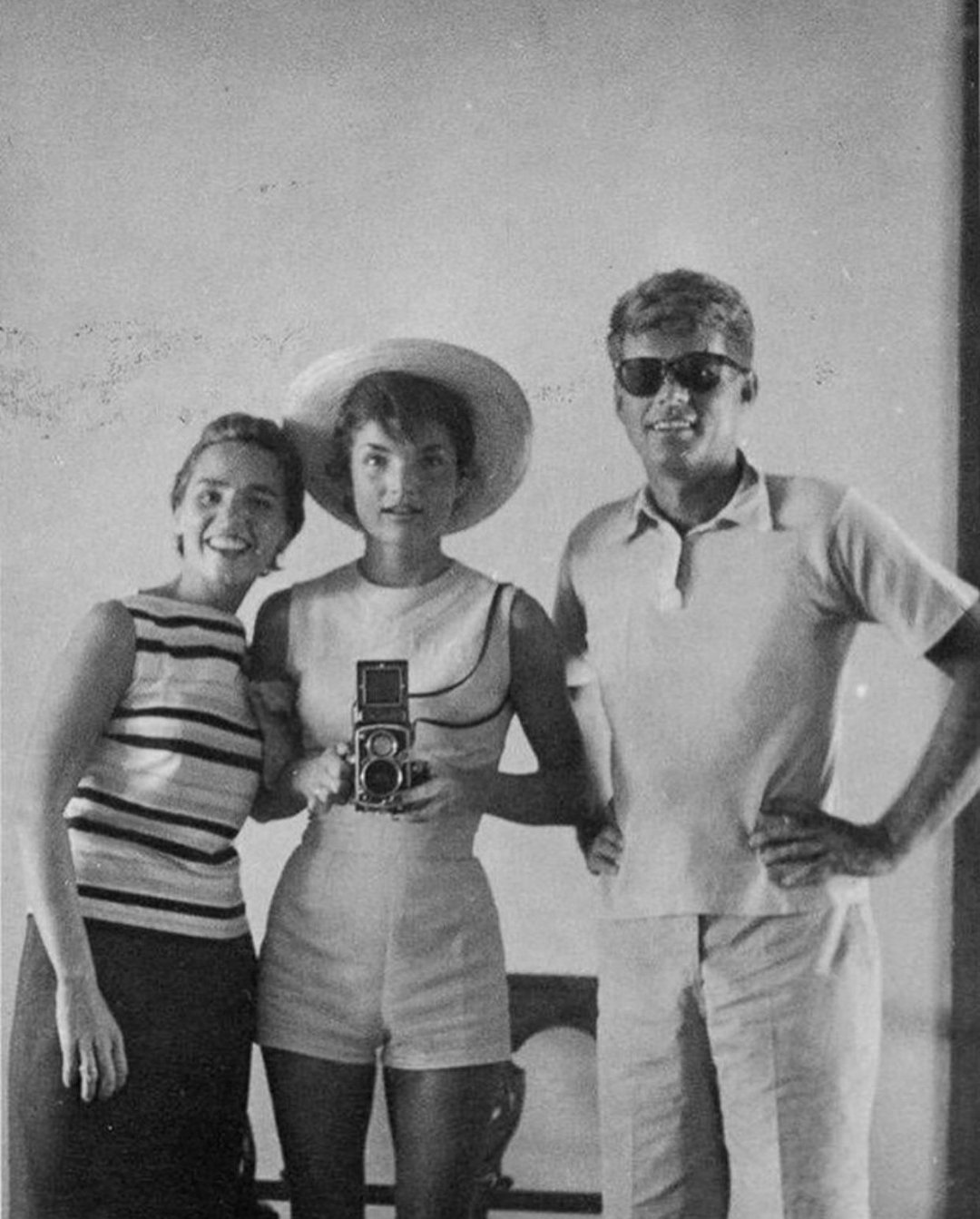 Three people pose for a black-and-white photo; one woman in a hat holds a camera, another woman smiles, and a man in sunglasses stands with his hand on his hip. All wear summer clothing and stand against a plain wall.