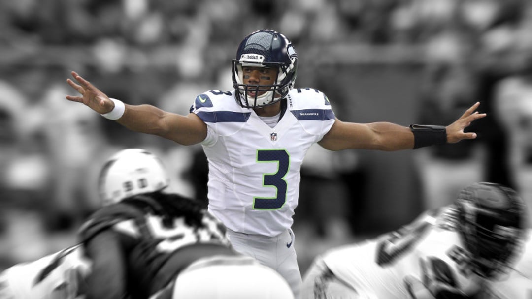 A football player in a white Seattle Seahawks uniform with the number 3 stands with arms outstretched, preparing to receive the snap, with opposing players blurred in the foreground.