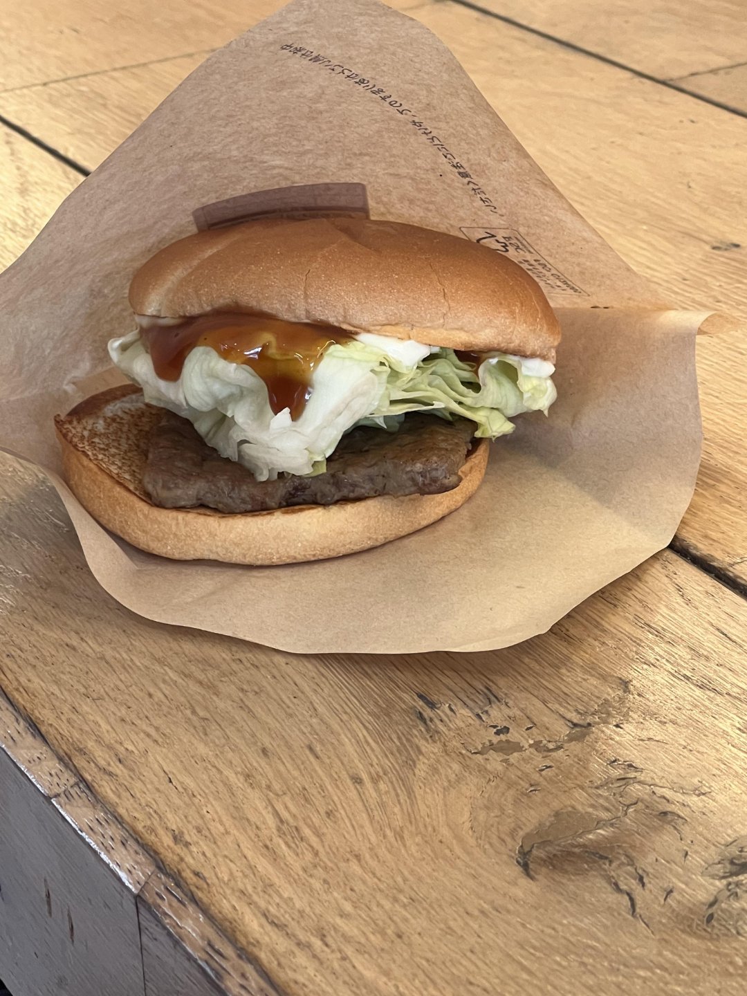 A hamburger with lettuce and sauce in a brown paper wrapper sits on a wooden table.
