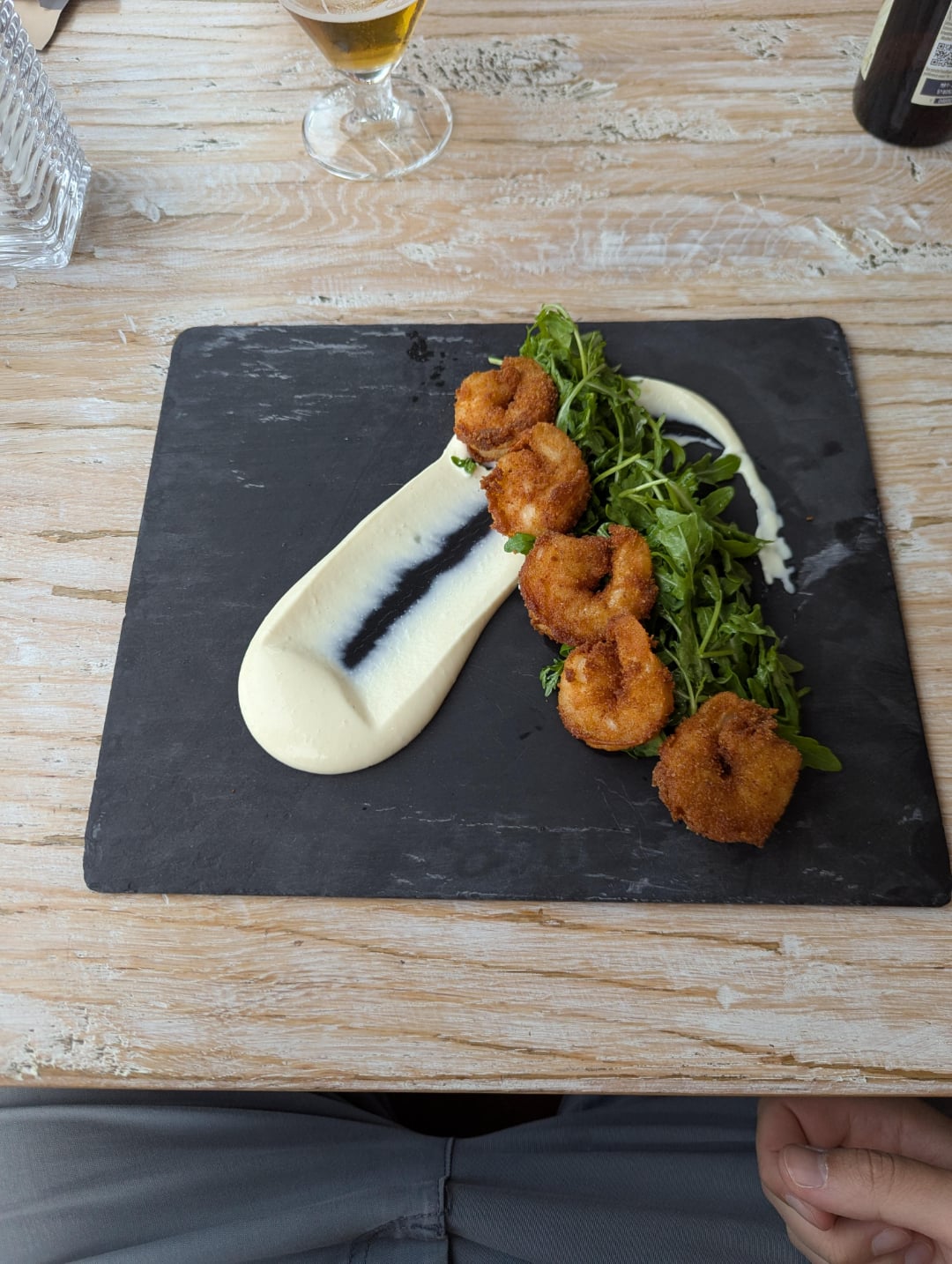 A black slate plate with five breaded shrimp, a swirl of creamy white sauce, a streak of dark sauce, and a small pile of arugula, on a light wooden table. Part of a person's legs and a drink are visible.