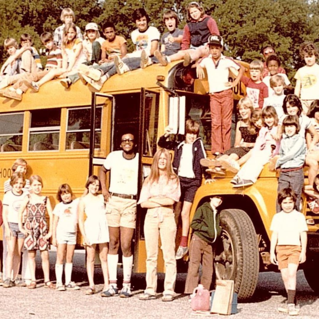A large group of children and a few adults pose on and around a yellow school bus, some sitting on the roof, steps, and hood. The scene appears to be from the 1970s based on their vintage clothing.