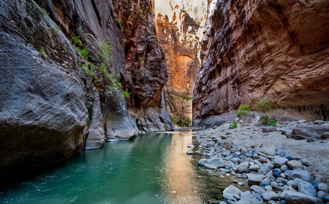 A river flows through a narrow canyon with steep, towering rock walls and scattered rocks along the shore. Sunlight illuminates part of the canyon, highlighting the rugged textures and vibrant colors.