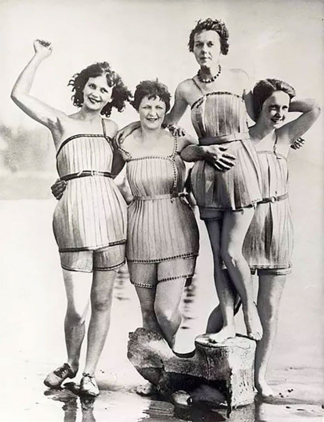 Four women in vintage swimsuits stand together at the beach, smiling and posing for the camera. One woman stands on a rock while the others stand beside her, all looking cheerful and relaxed. The image is in black and white.
