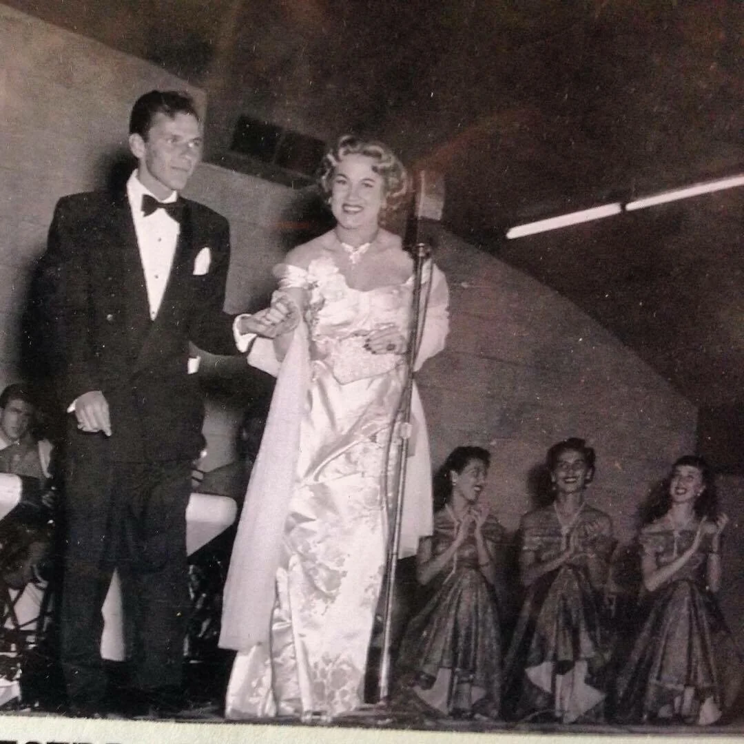 A man in a tuxedo and a woman in an elegant gown stand on stage holding hands by a microphone, while four women in matching dresses sit behind them, clapping and smiling. The setting has a vintage, black-and-white feel.
