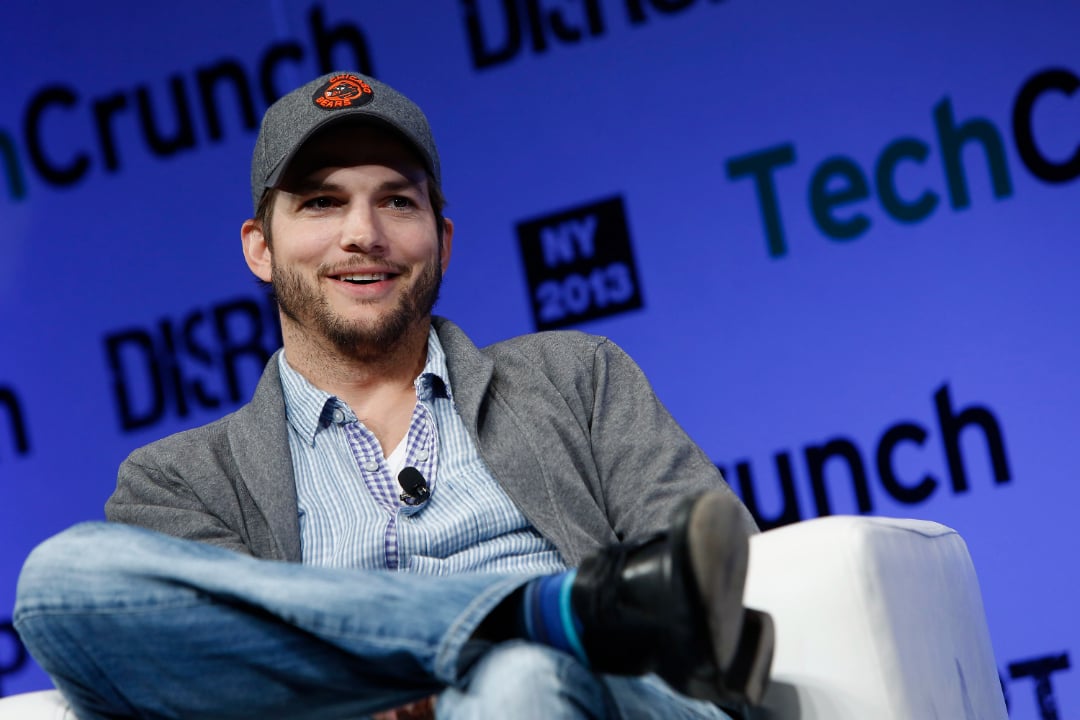 A man wearing a gray jacket, blue shirt, jeans, and a baseball cap sits cross-legged on a white chair, smiling, against a backdrop with TechCrunch and Disrupt NY 2013 logos.