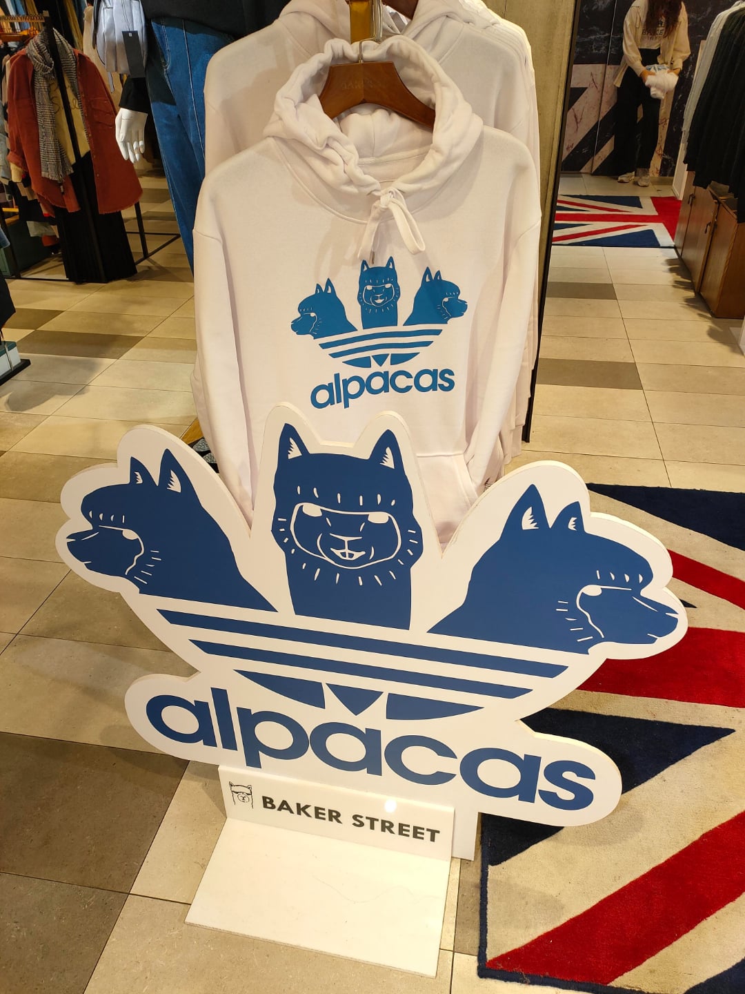A white hoodie with a blue graphic of a smiling alpaca and the word "alpacas" hangs on display. In front, a matching stand shows three cartoon alpaca faces and "alpacas" above a "Baker Street" label.