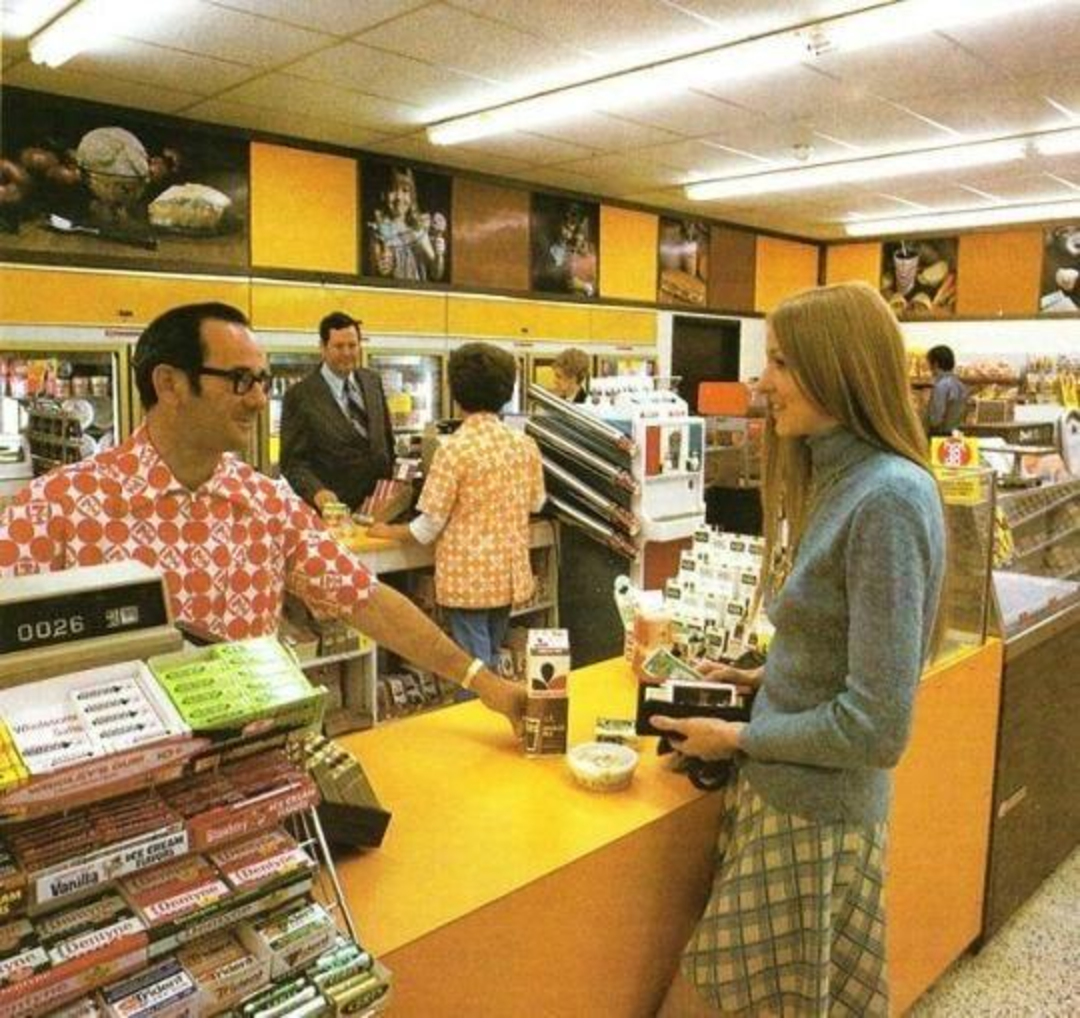 A woman in a blue sweater and plaid skirt talks to a cashier at a brightly lit 1970s grocery store. Shelves of candy, gum, and other goods are on the counter. Other shoppers and employees are seen in the background.
