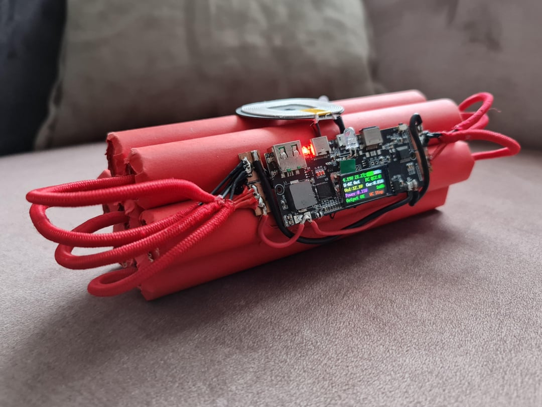 A homemade electronic device with red cylinders, resembling dynamite sticks, is connected by wires and a digital timer with a display is attached, resting on a beige surface.