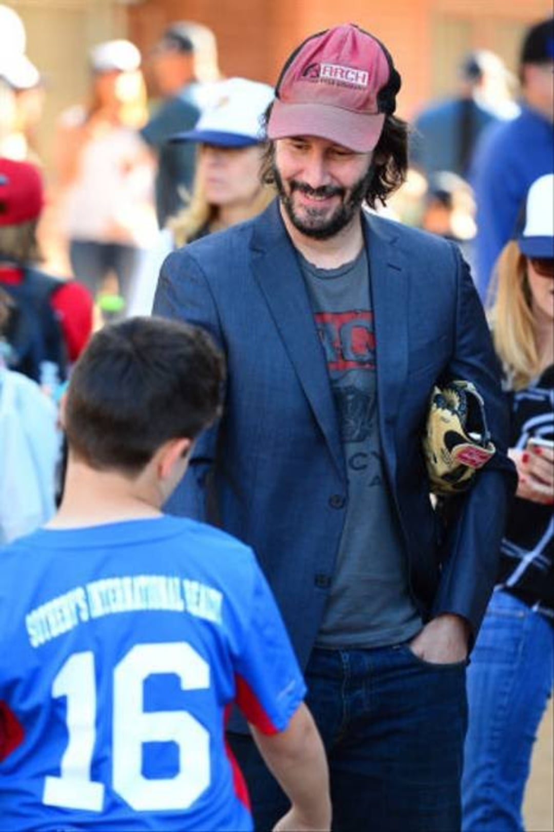 A man in a red cap and blazer smiles while talking to a boy in a blue sports jersey with the number 16 on the back, surrounded by other people in casual clothes outdoors.