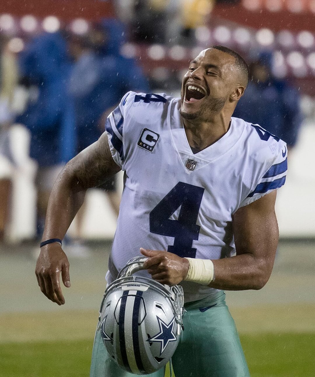A football player in a Dallas Cowboys uniform, wearing number 4, laughs while holding his helmet on a rainy football field.