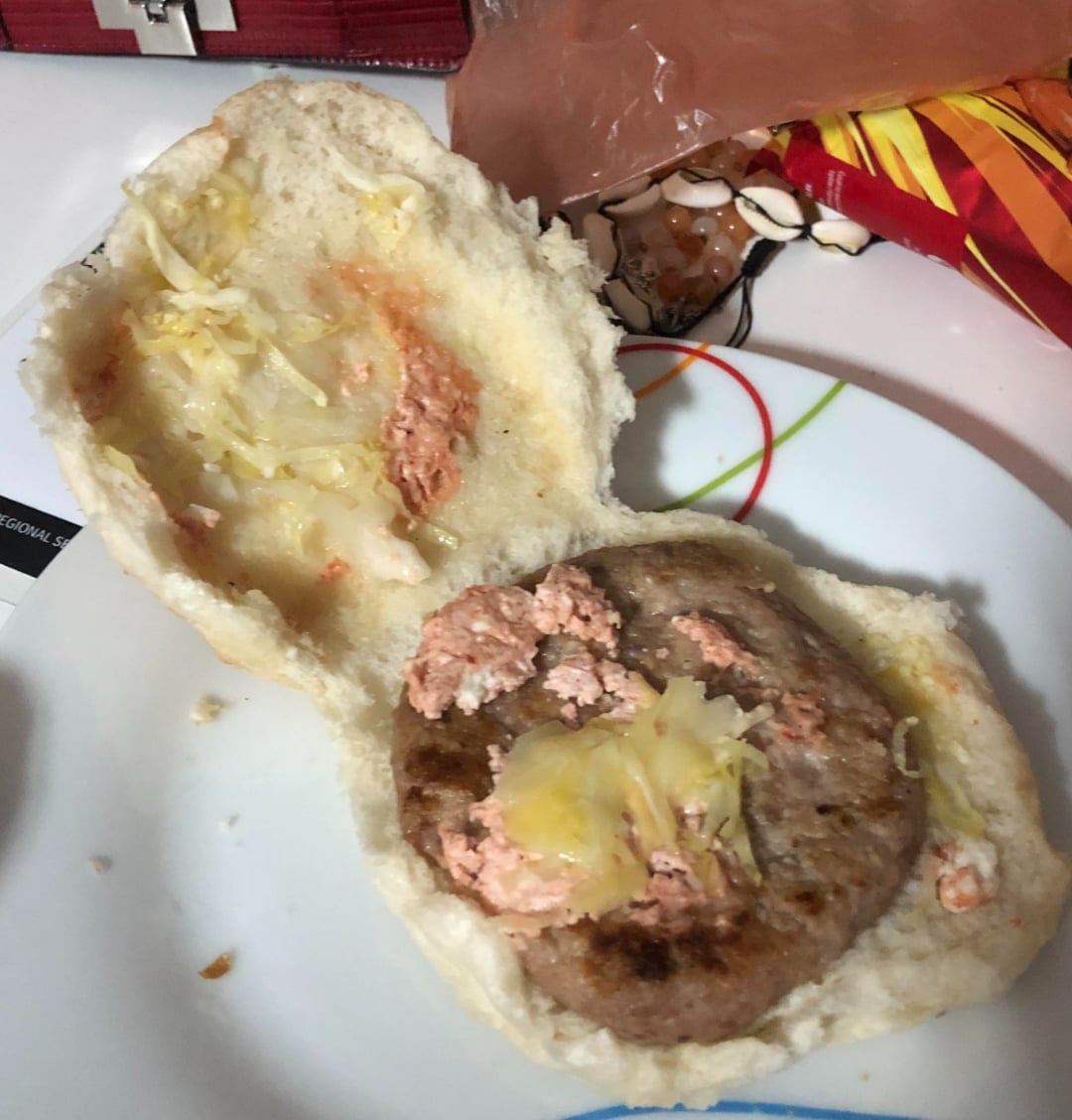 An open hamburger bun on a plate with a cooked patty, some shredded cheese, and a spread that looks unevenly applied. The background shows a plastic bag and a packet with a colorful design.