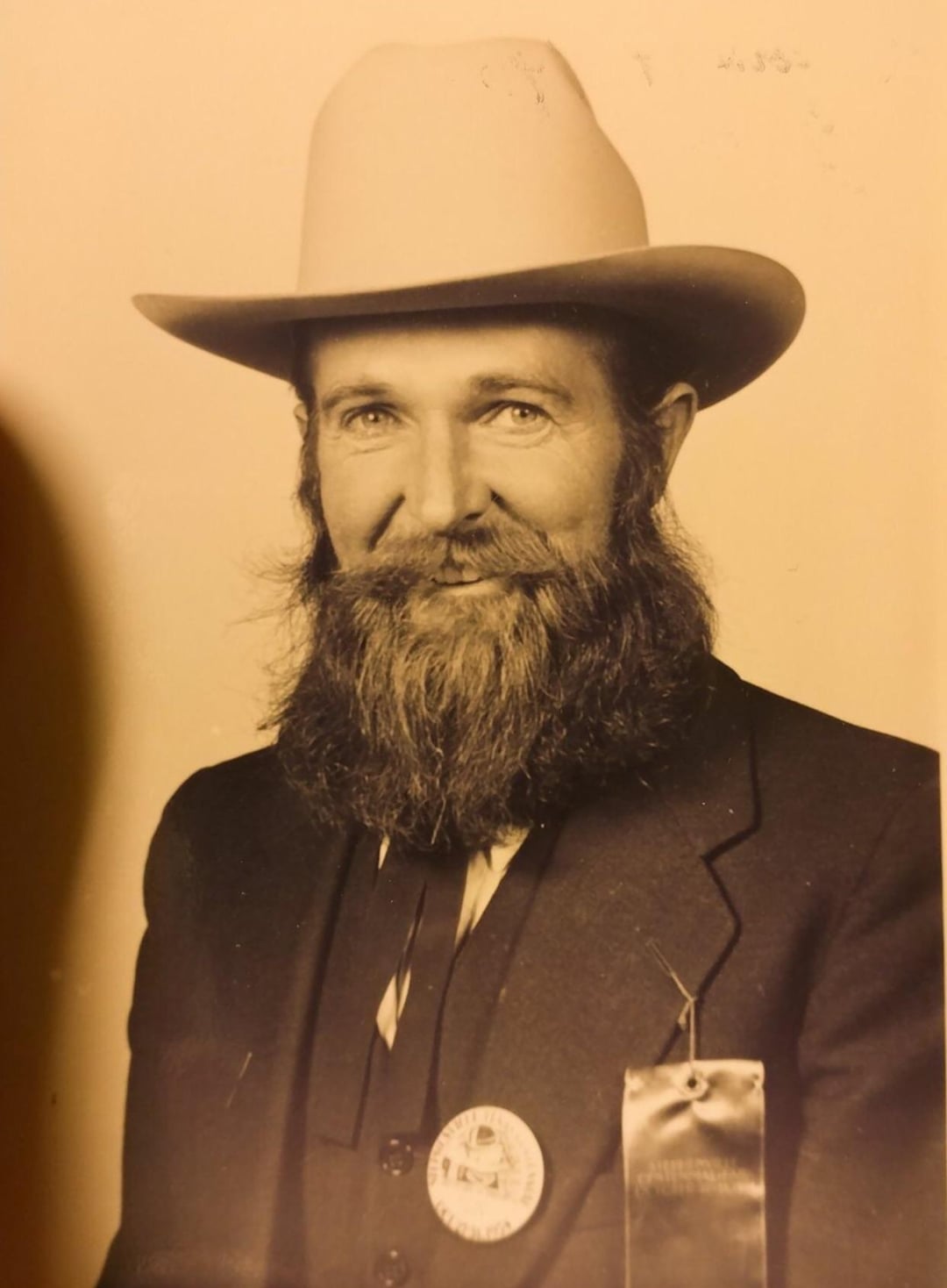 Sepia-toned portrait of a man with a full beard, wearing a wide-brimmed cowboy hat, suit, and bolo tie. He has a badge and ribbon pinned to his jacket, and he is looking at the camera with a slight smile.