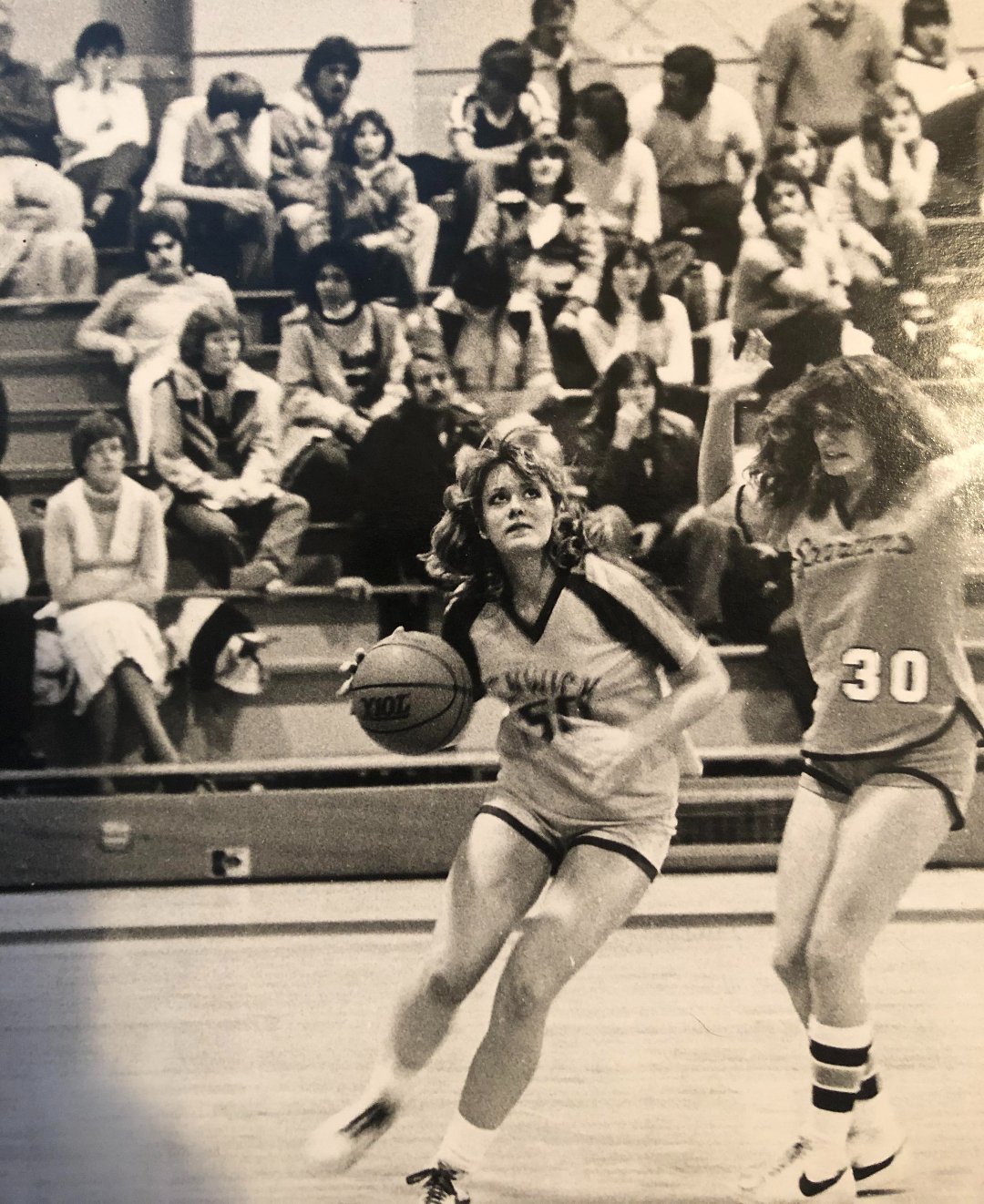 A vintage black-and-white photo shows a girls’ basketball game. A player in a "Savick" jersey dribbles the ball, while a defender in a "30" jersey follows closely. Spectators watch from the bleachers in the background.