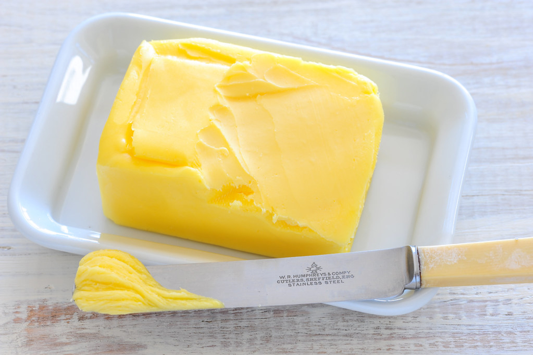 A rectangular block of yellow butter sits on a white dish, with a butter knife resting in front. The knife has a small amount of butter on its blade. The scene is set on a light-colored wooden surface.
