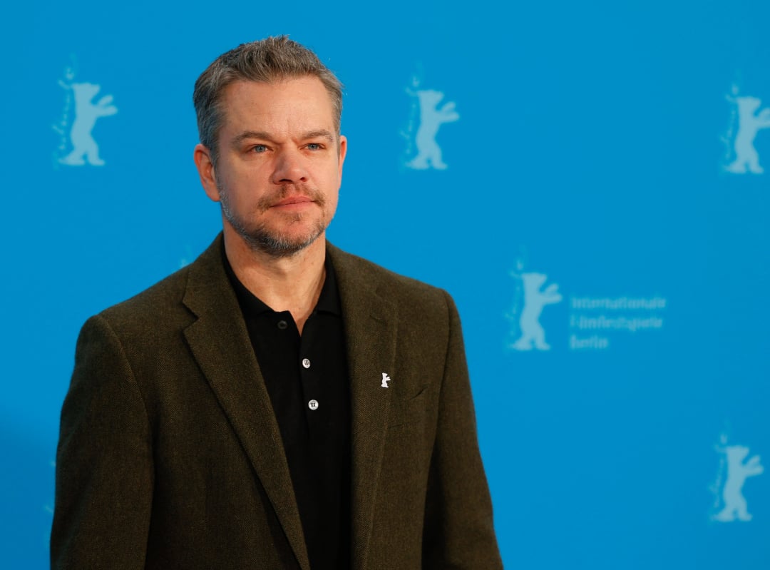 A man with short gray hair and a beard stands in front of a blue background with white bear logos and text. He wears a dark green blazer over a black buttoned shirt and looks slightly to the left.