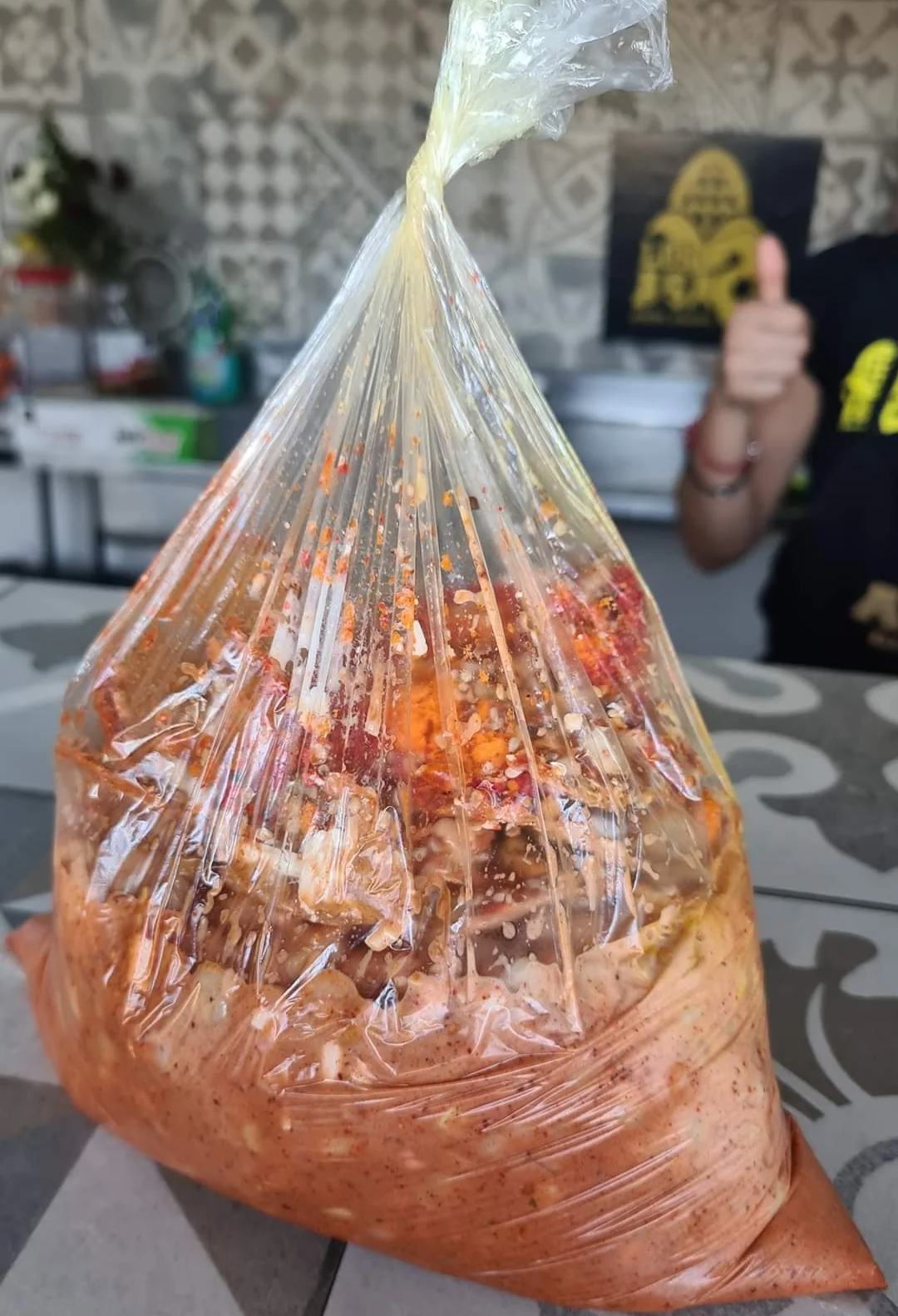 A clear plastic bag filled with a spicy, reddish marinated food mixture sits on a counter. In the background, a person gives a thumbs-up, with tiles and kitchen items visible.