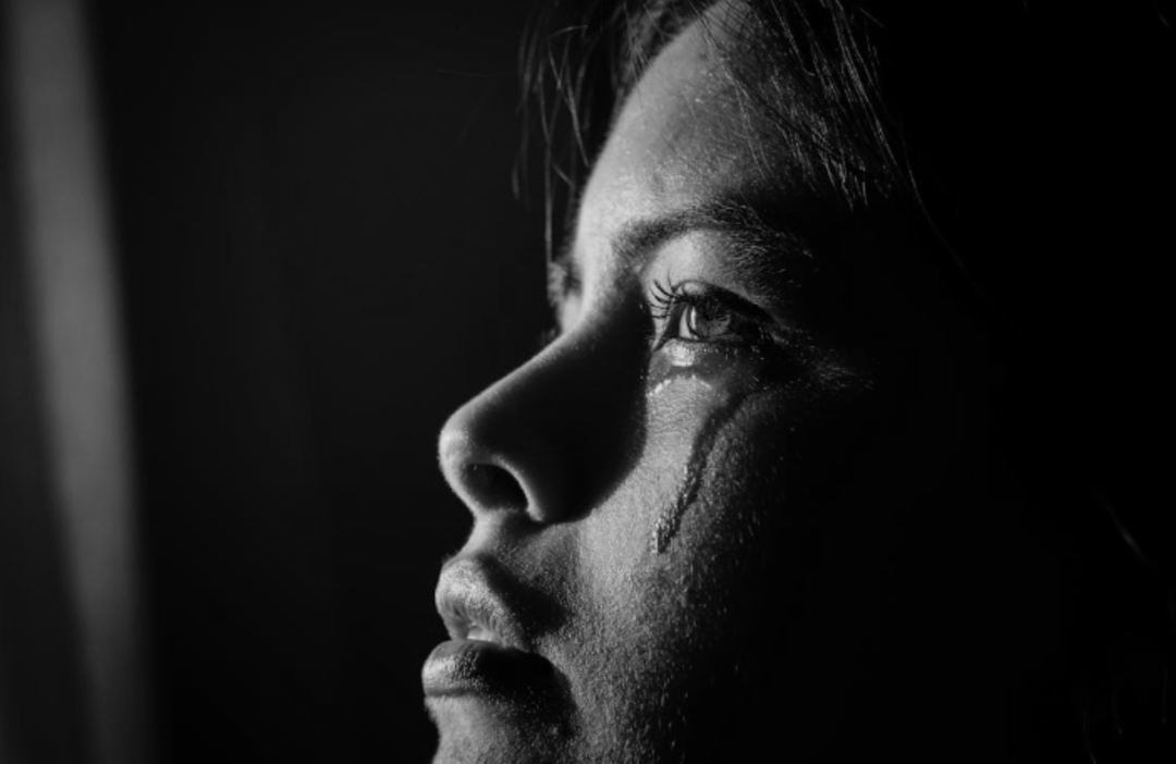 Close-up black and white photo of a person's face in profile, looking slightly upward, with a tear running down their cheek, conveying a sense of sadness or contemplation.