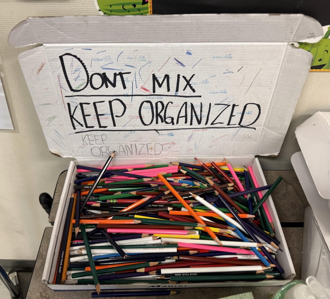 An open box filled with assorted colored pencils. The inside lid has a handwritten message: "DON'T MIX, KEEP ORGANIZED," surrounded by scribbles in different colors.