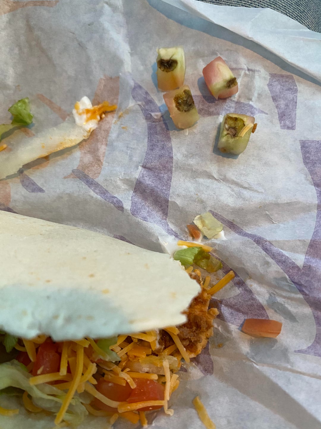 A partially eaten taco sits on a wrapper, with a few moldy diced tomatoes and other taco ingredients scattered nearby.
