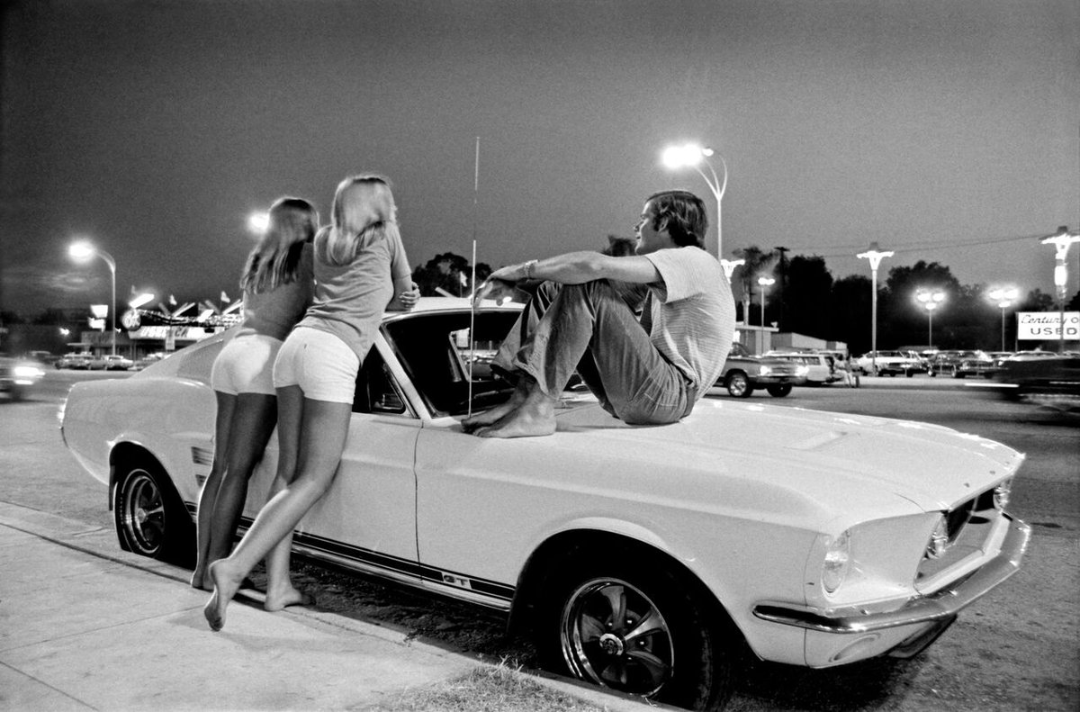 Three young people, two women standing and a man sitting barefoot, hang out on the hood of a classic white car at night in a lit-up parking lot, with neon signs and parked cars in the background.