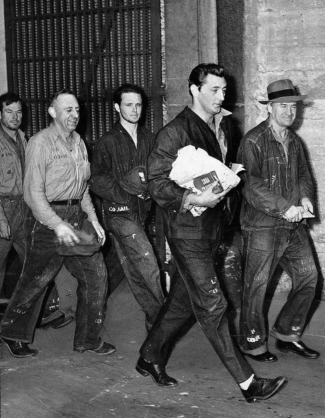 A group of men in dark prison uniforms, some smiling, walk together indoors. One man in front carries clothes and a book. The background shows a barred door and a concrete wall.