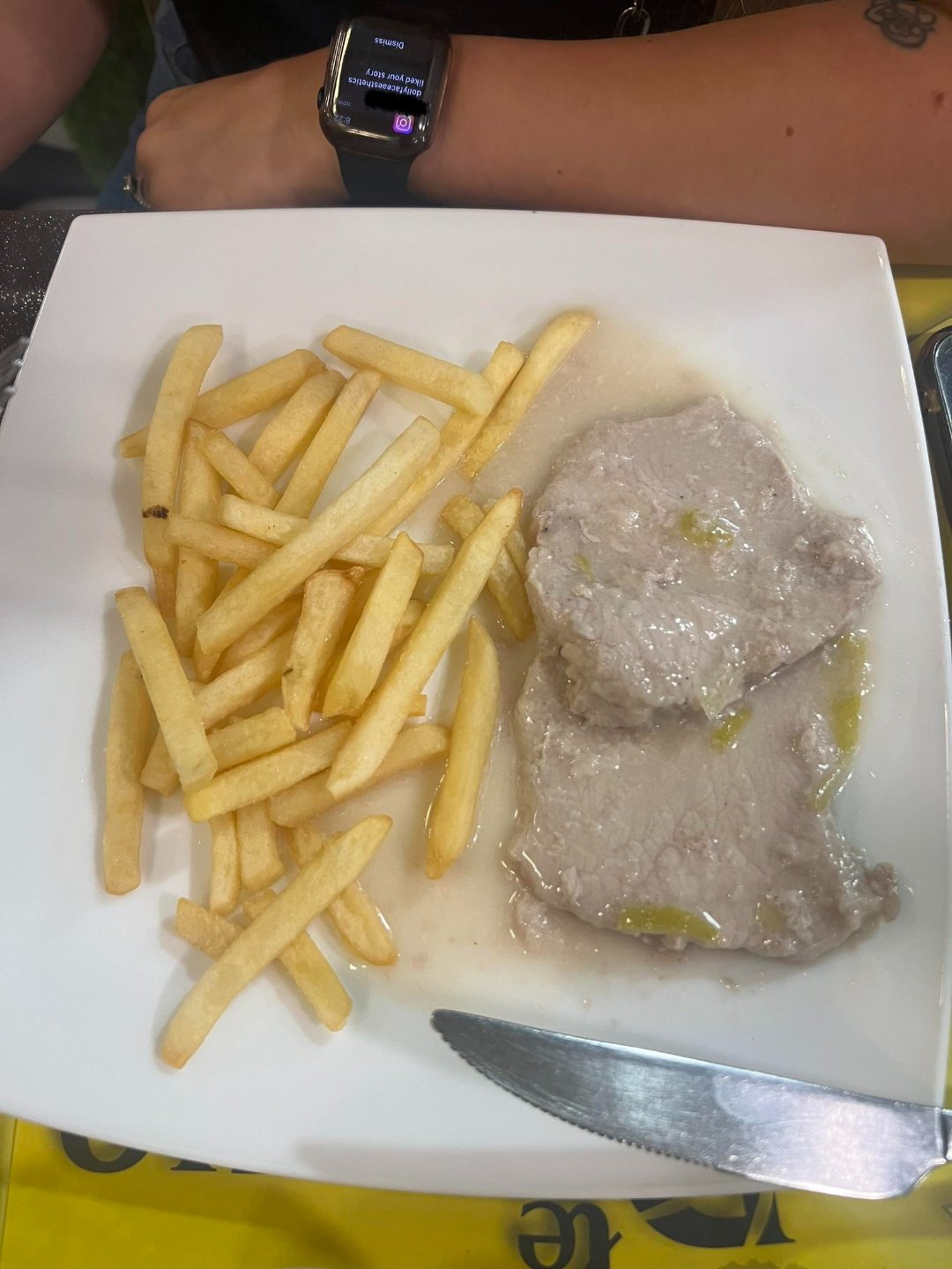 A square white plate with French fries and two pieces of meat in a light sauce. A knife rests on the plate. In the background, a person with a smartwatch and a tattoo is seated at the table.