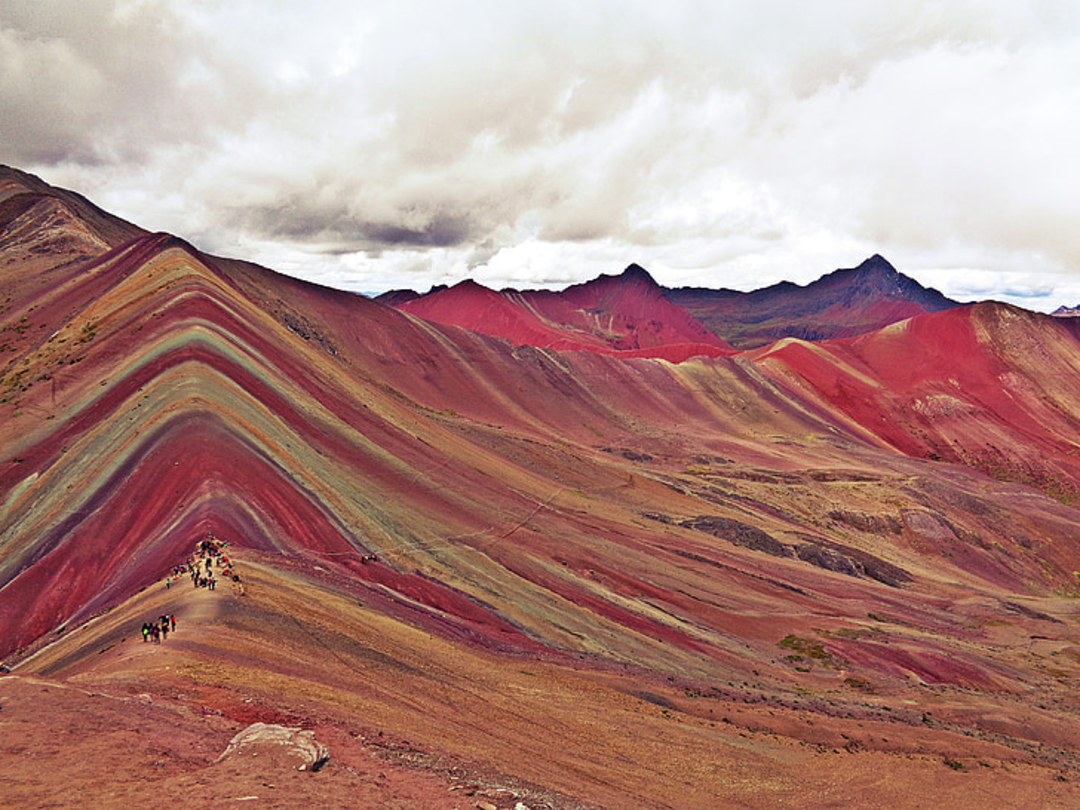 A group of hikers walk along a trail on Rainbow Mountain in Peru, featuring colorful, layered stripes of red, yellow, and green earth under a cloudy sky with distant dark mountains.