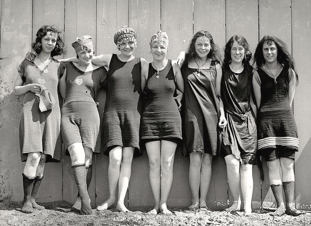 Seven women in vintage swimsuits stand side by side, smiling and posing in front of a wooden wall. The image appears to be black and white and from the early 20th century.