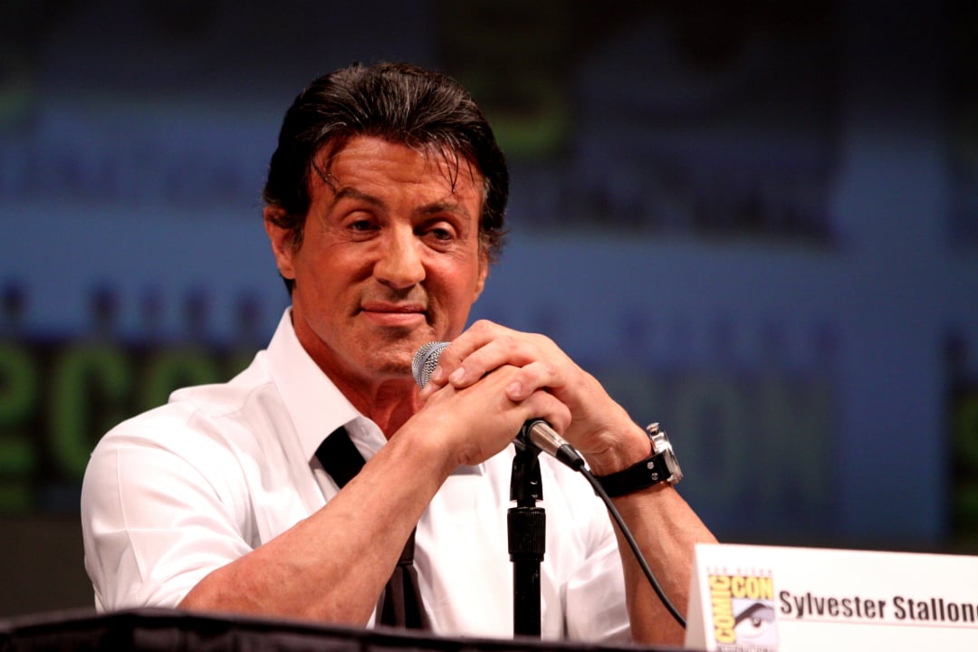 A man in a white shirt and loosened tie sits at a panel table, holding a microphone and smiling. A Comic-Con placard with the name "Sylvester Stallone" is visible in front of him.