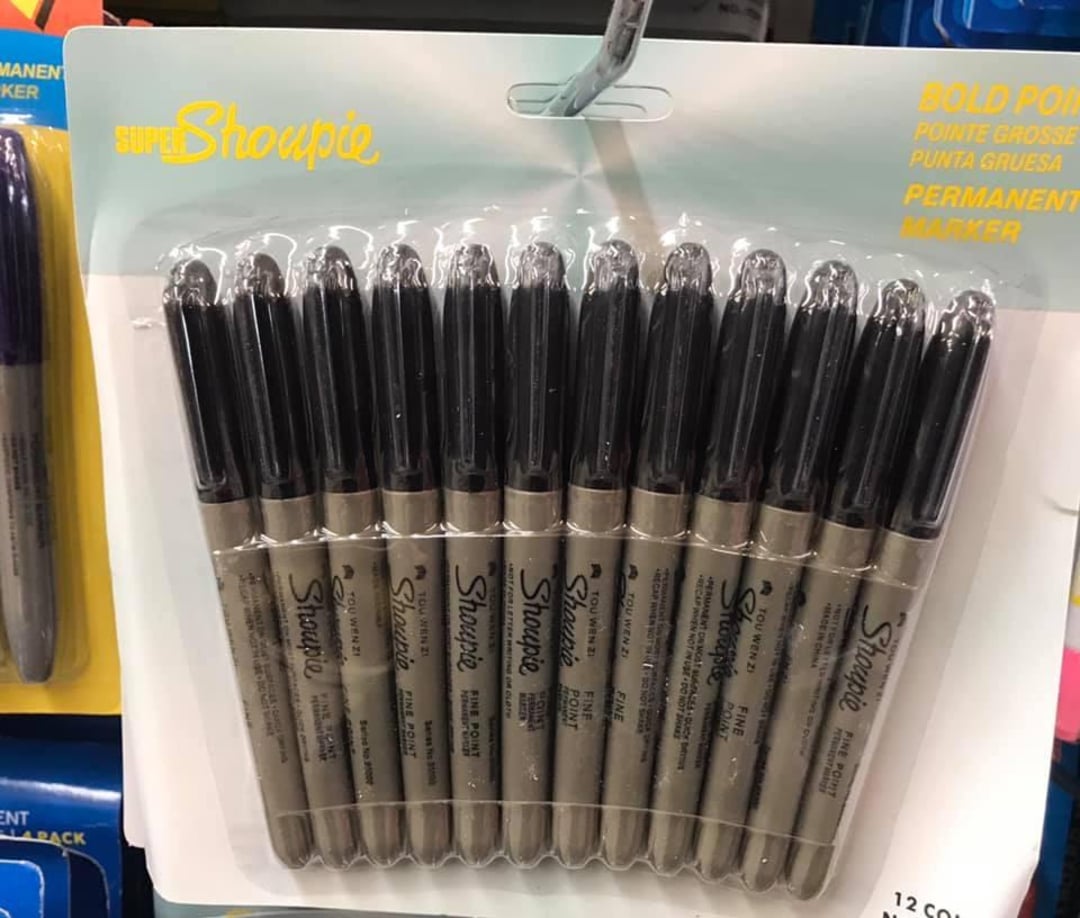 A package of 12 black Super Sharpie permanent markers is displayed on a store shelf, with the markers arranged side by side in clear plastic packaging and labeled "Bold Point.