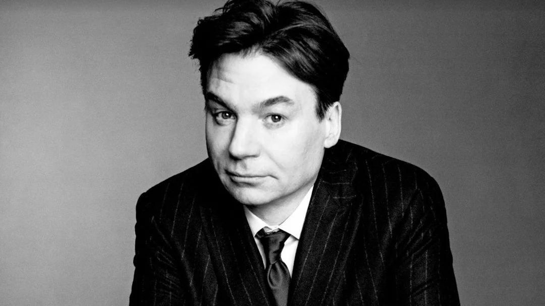 A man in a dark pinstripe suit and tie looks at the camera with a neutral expression. The background is plain and gray, and the image is in black and white.