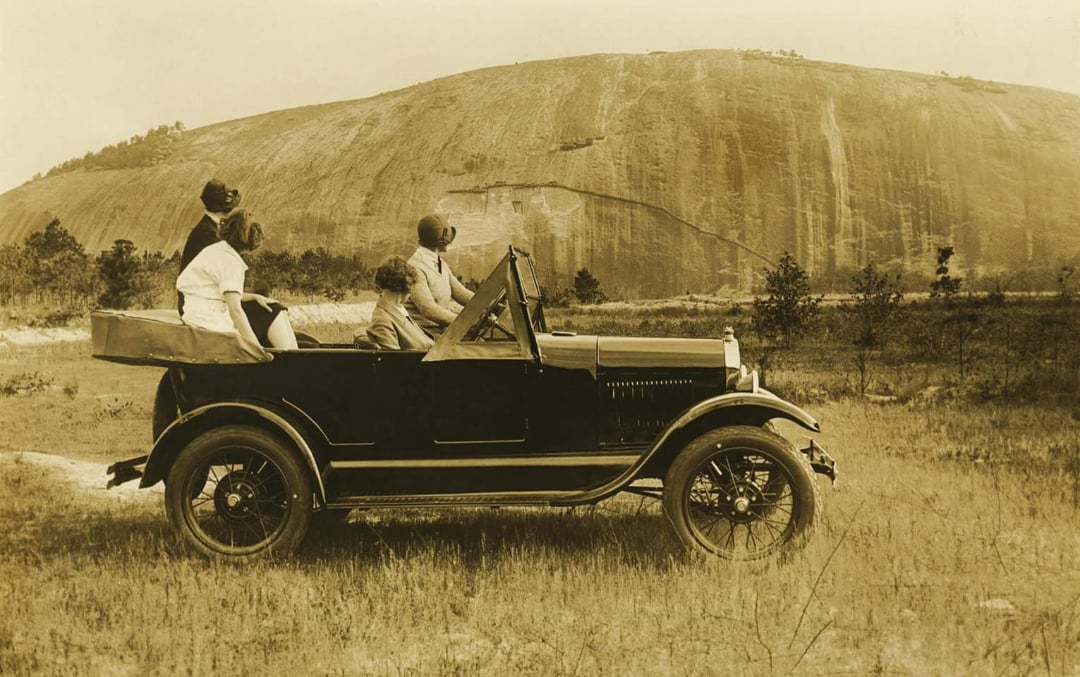 Four people sit in a vintage open-top car parked on grass, looking toward a large rocky hill in the background under a clear sky. The image appears to be from the early 20th century.