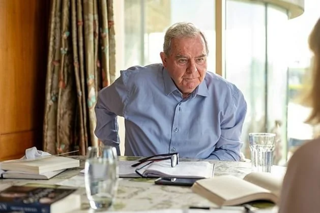 An older man in a blue shirt sits at a table with books, papers, and glasses of water, looking thoughtfully at someone out of view. Sunlight streams in from large windows behind him.