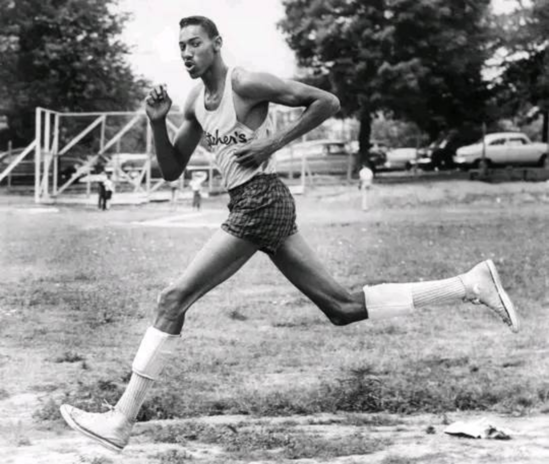 A tall male athlete in a tank top, shorts, and high socks is running outdoors on a dirt path, with trees, parked cars, and playground equipment visible in the background.