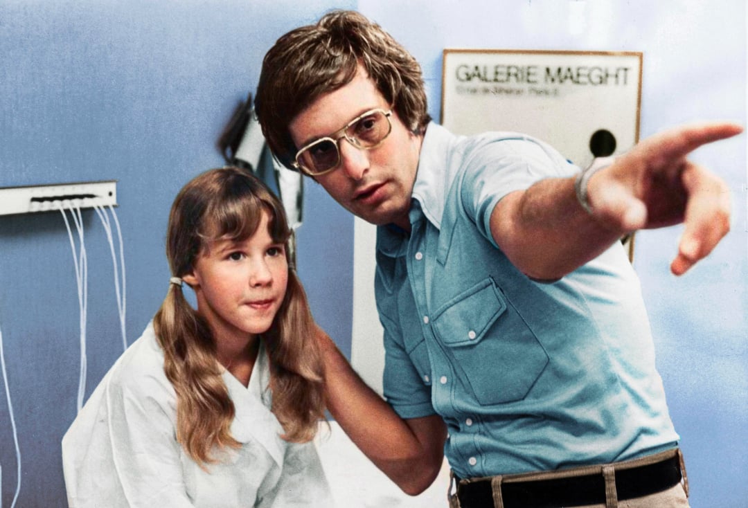 A man in glasses and a blue shirt points to something off-camera while a young girl with long hair in pigtails sits next to him, wearing a white robe. They are indoors with medical wires and a "Galerie Maeght" sign behind them.