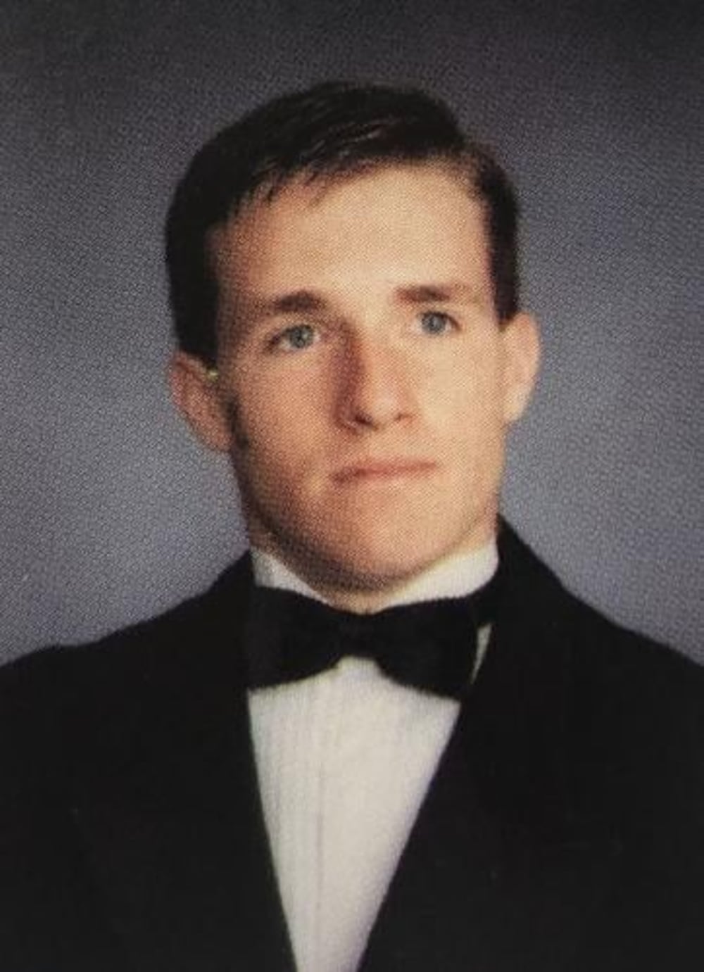A young man with short brown hair wearing a black tuxedo, white dress shirt, and black bow tie poses for a formal studio portrait against a gray background.