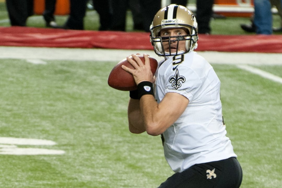 A football player wearing a white New Orleans Saints jersey with the number 9 prepares to throw a football during a game on a turf field.