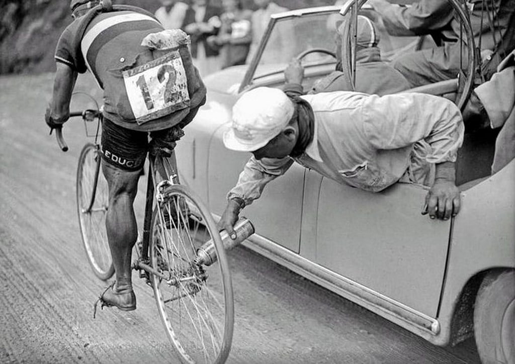 A cyclist is handed a water bottle by a man leaning out of a car window during a race. The cyclist wears a number 12 on his back, and the scene appears vintage, with spectators blurred in the background.