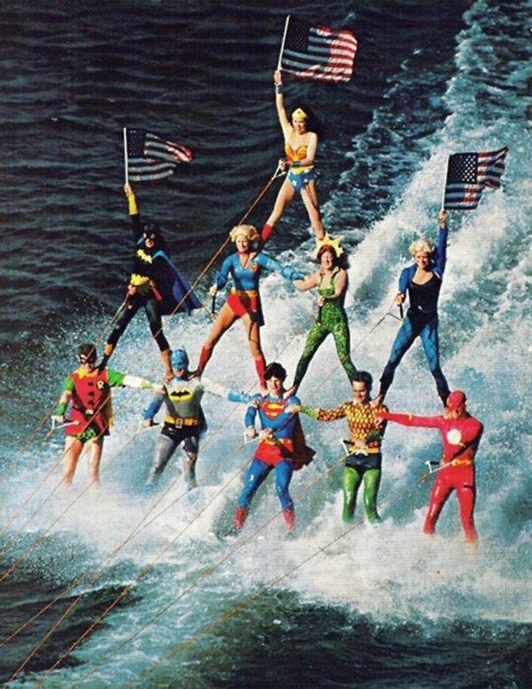 A group of people in colorful superhero costumes form a human pyramid while water skiing, holding American flags above the water’s surface. The scene is vibrant and dynamic with water spray around them.