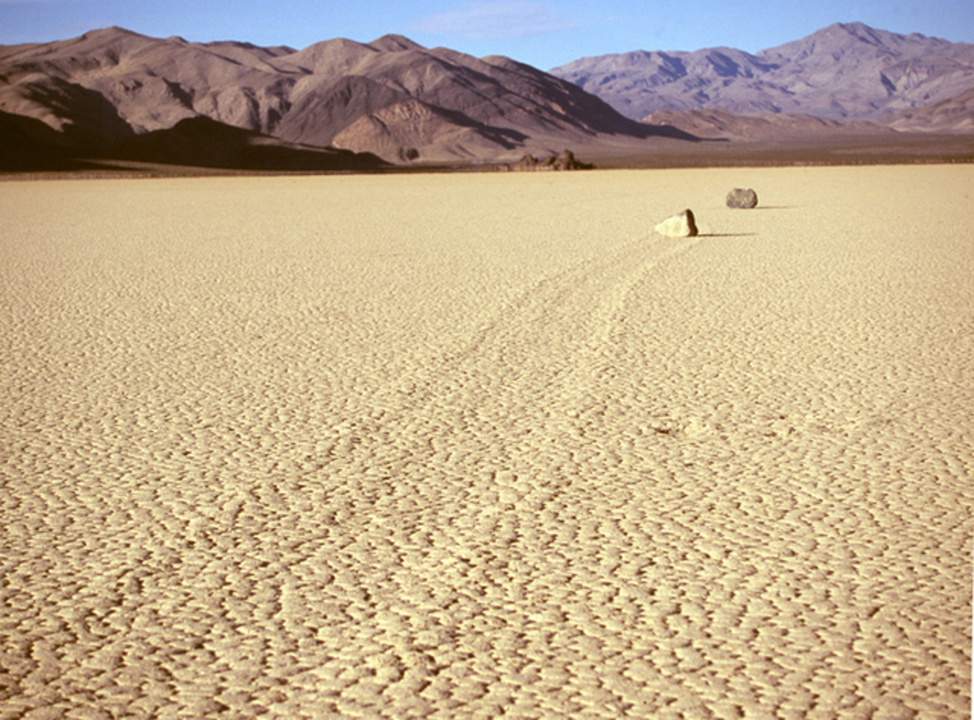 Two rocks sit on the cracked, dry surface of a desert playa, with long trails behind them suggesting movement. Mountains rise in the background under a clear blue sky.