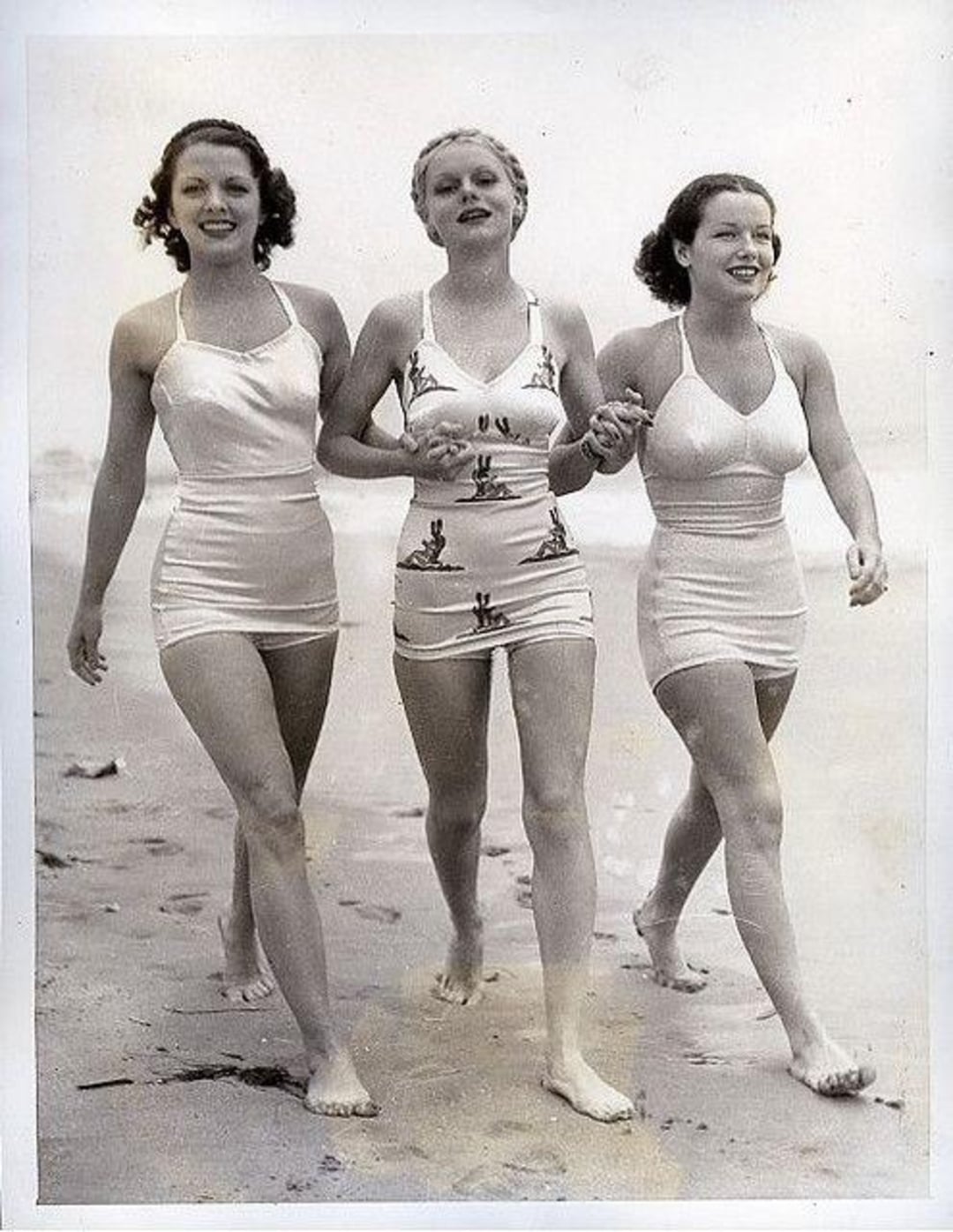 Three women in vintage swimsuits walk barefoot on a sandy beach, holding hands and smiling. The photo is black and white, giving it a nostalgic, early 20th-century feel.