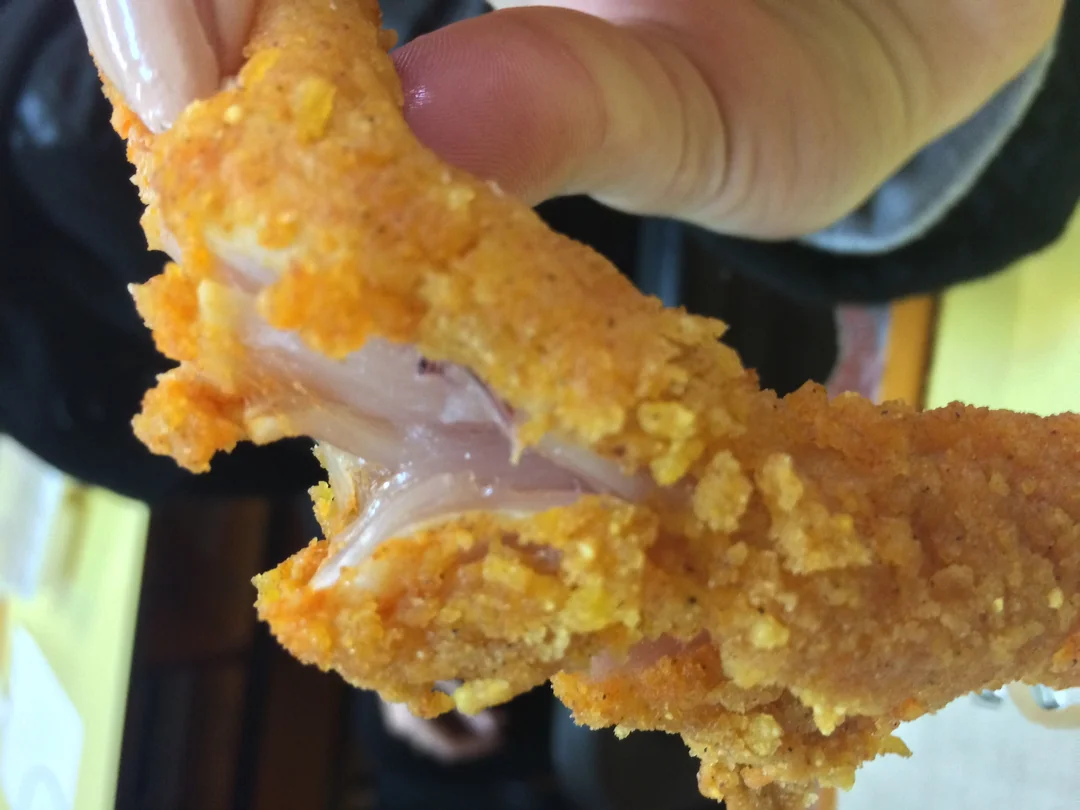 A hand holding a piece of fried chicken with a crispy, golden-brown coating, revealing uncooked, pink meat inside.