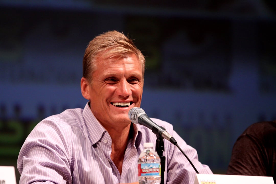 A smiling man with short blond hair sits at a panel, speaking into a microphone. He wears a light purple shirt and there is a water bottle in front of him. The background is blurred with indistinct text and logos.
