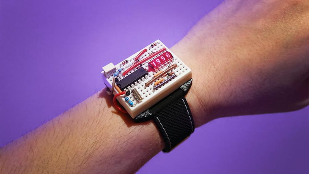 A wrist wearing a homemade digital watch with exposed electronic components and a red LED display, mounted on a black strap, against a purple background.