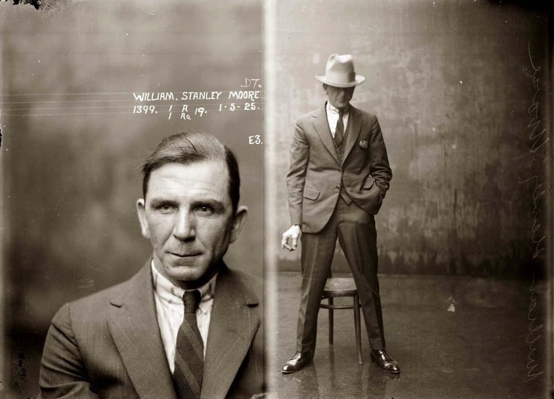 A vintage mugshot shows a man in a suit and tie. The left side is a close-up of his face, while the right side shows him standing, holding a cigarette, wearing a fedora, and posing beside a chair. Writing appears next to his face.