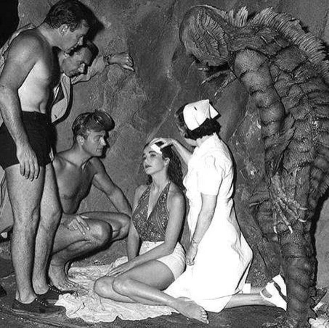 A black-and-white photo shows four men, one in a creature costume, and a woman in a nurse outfit attending to a seated woman. The group appears to be on a film set, possibly near a rocky backdrop.