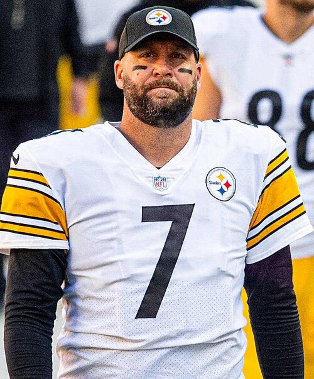 A football player wearing a white Pittsburgh Steelers jersey with the number 7, a black cap, and black eye paint stands on the field. Other teammates in similar uniforms are visible in the background.